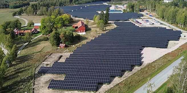 Flygfoto över Better Energy Swedens solpark i Studsvik, Nyköping. Stora fält täckta av solpaneler breder ut sig intill en skog och några röda lantgårdsbyggnader. Solpanelerna följer markens form och sträcker sig bort mot horisonten. Till höger syns en mindre väg och parkerade bilar. Solparken är Sveriges största med en yta på cirka 23 hektar.