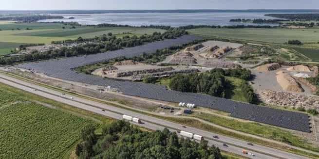 Bilden visar Ekens solcellspark med solpaneler utplacerade i ett oregelbundet mönster kring en gammal grustäkt. I förgrunden syns en större väg med passerande fordon, och i bakgrunden breder odlingslandskap och en sjö ut sig. Panelerna är anpassade efter terrängen och delvis omgivna av träd och högar med grus eller sten. Bilden är tagen från ovan under klart väder och visar hur solparken utnyttjar mark som annars inte används för jordbruk.