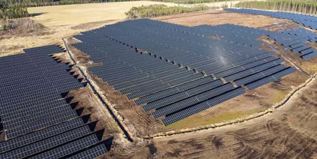 Bilden visar Henja solcellspark med ett stort antal solpaneler utplacerade i långa rader över en röjd skogsmark. Marken är delvis bar och omgiven av träd och öppna fält. Solpanelerna följer markens lutning och skapar ett rutmönster. Inga stängsel eller barriärer syns runt anläggningen. Bilden är tagen från ovan under klart väder och visar hur solparken integrerats i det naturliga landskapet.