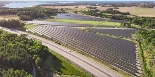 Bilden visar Strängnäs solcellspark med stora fält täckta av solpaneler i ett öppet landskap. I förgrunden syns en väg som löper längs kanten av solparken. Solpanelerna är placerade i långa rader med gröna områden mellan fälten. I bakgrunden syns skog, åkermark och en glimt av vatten. Bilden är tagen från ovan i klart väder, vilket ger en god överblick över parkens utbredning.